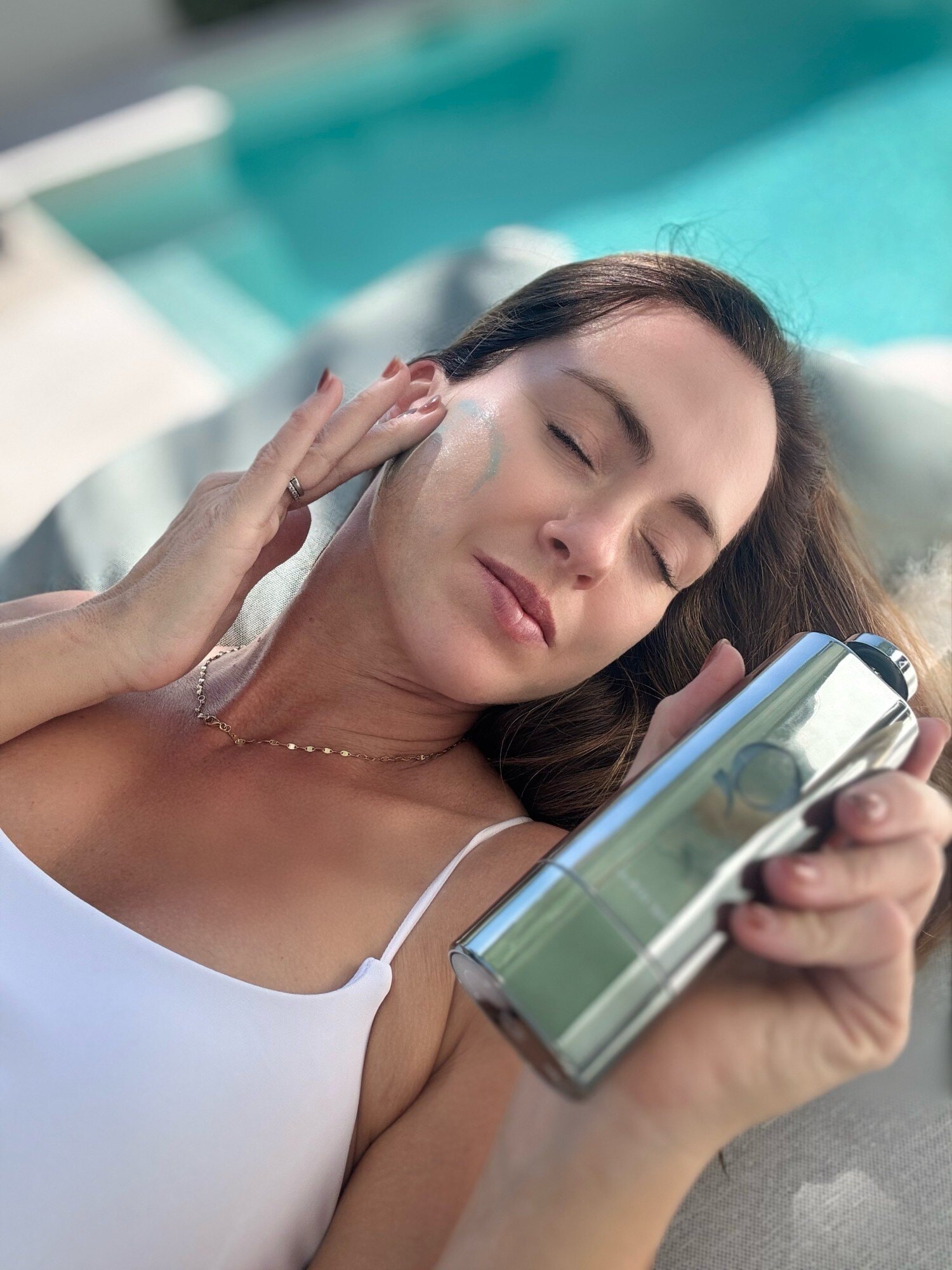 Sigourney Cantelo lying poolside with a streak of blue serum on her cheek, holding the Qure Blue+ Face Serum