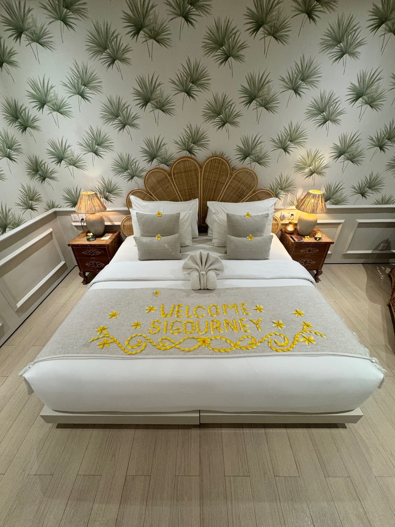 Welcome Sigourney spelled in frangipani petals across the bed of a goddess room at Escape Haven, under a rattan headboard and palm frond wallpaper