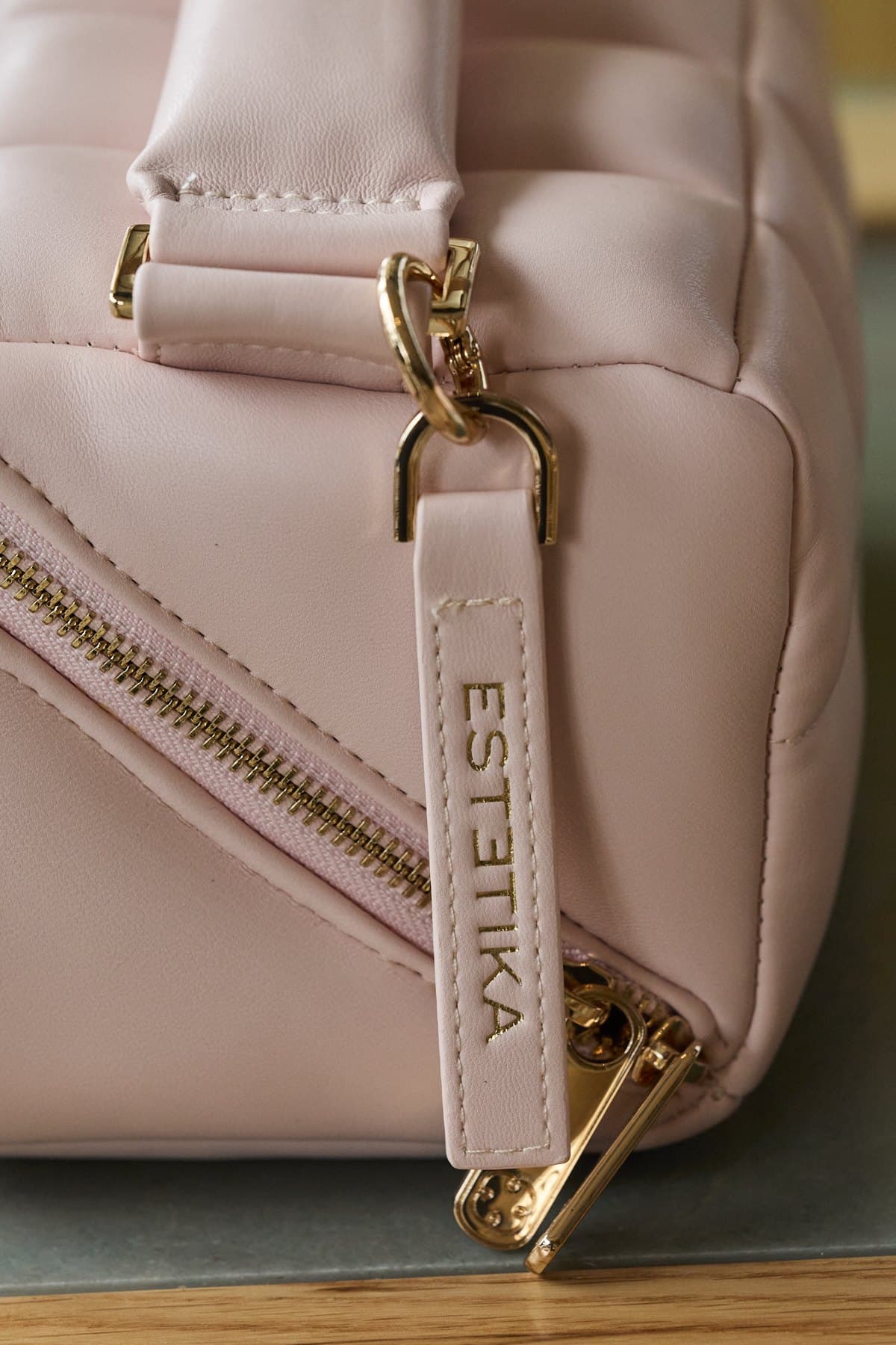 A pink quilted Estetika makeup bag with its leather zip pull, on a green marble table