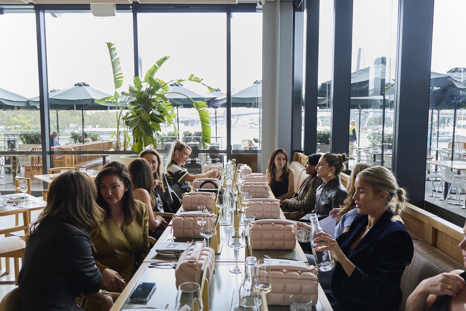 Guests seated along the long table at Lua, pink bags set at each place and harbour windows behind