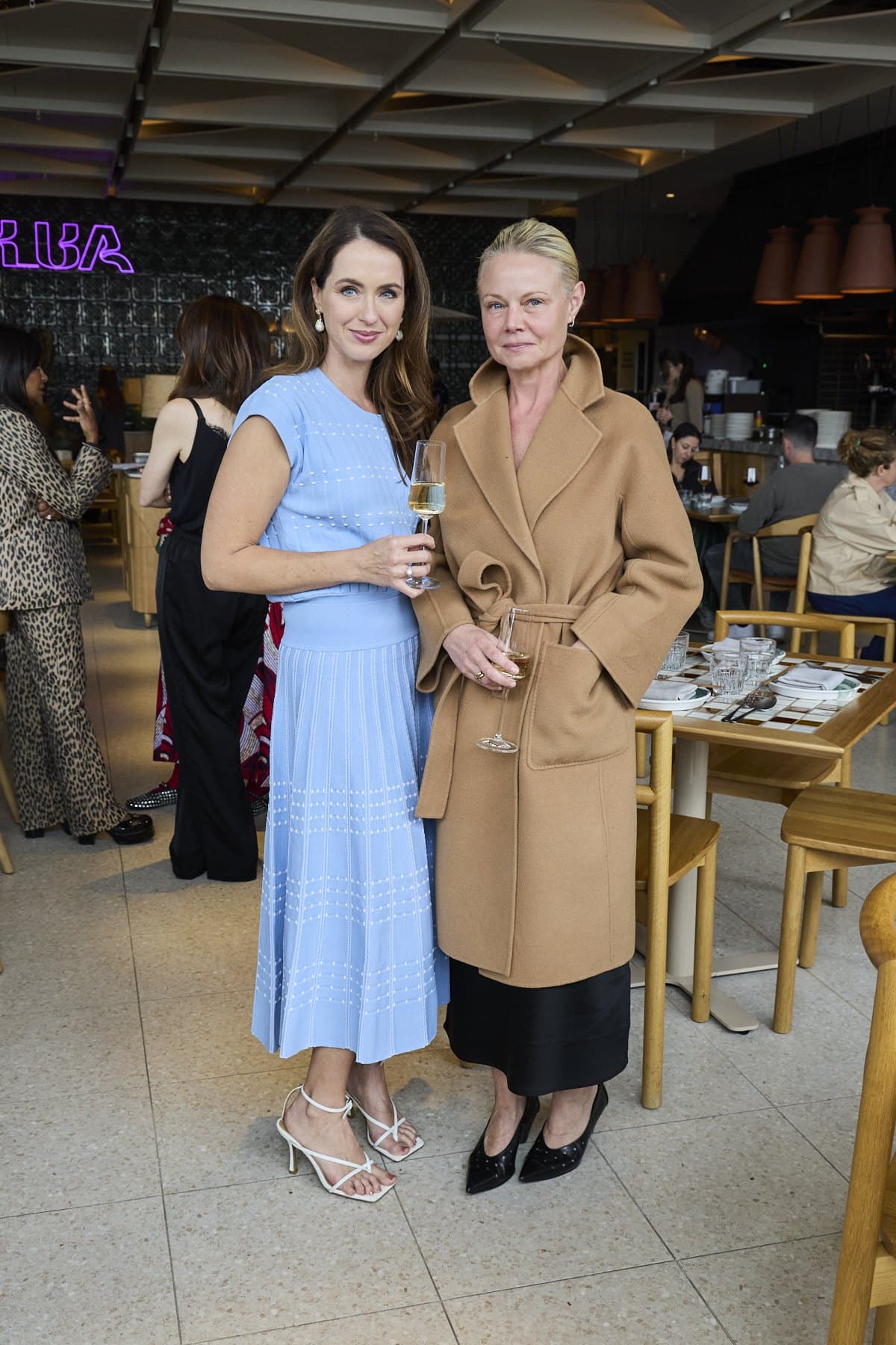 Sigourney in her blue dress standing with a guest in a camel wrap coat at Lua