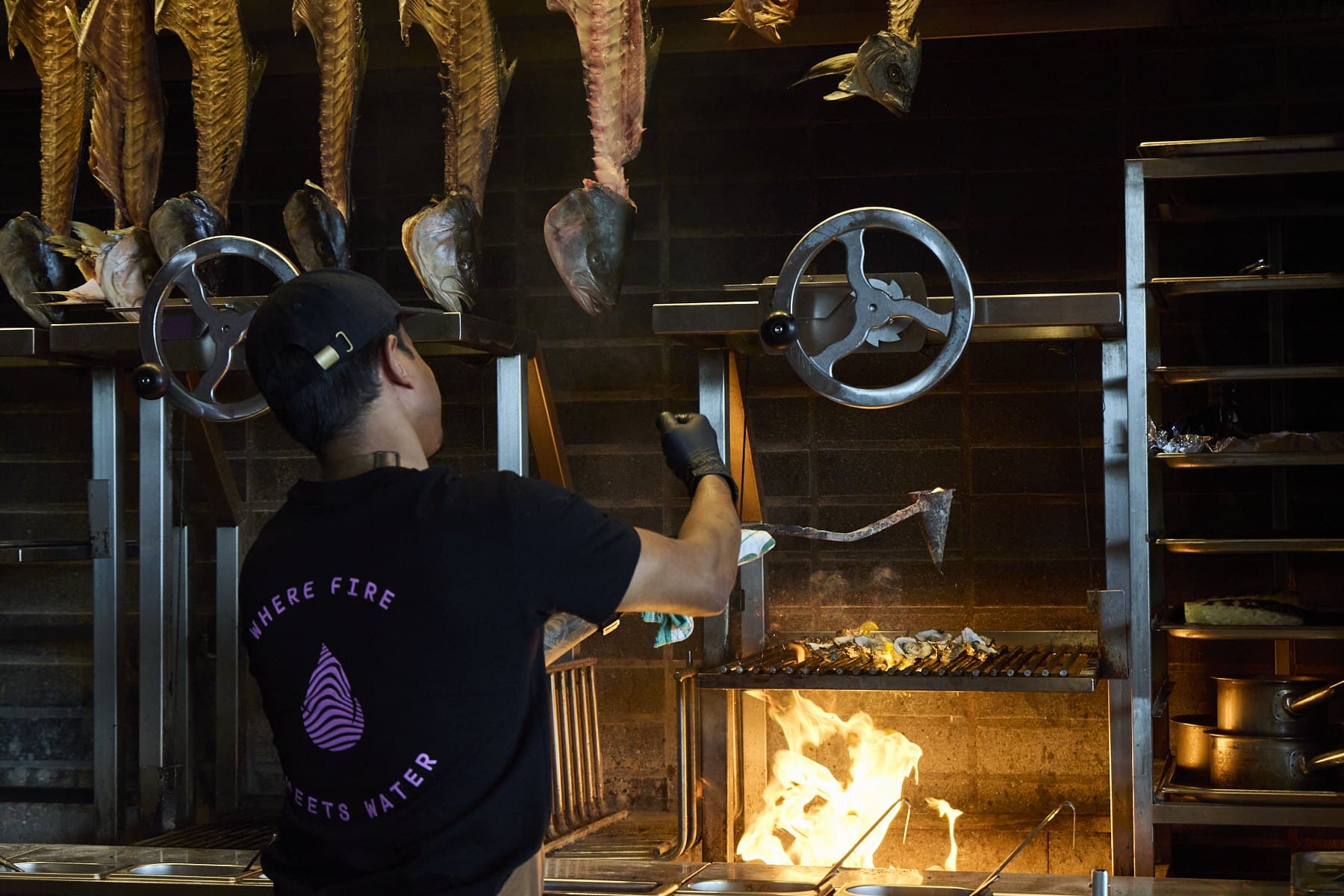 A chef at Lua's charcoal hearth working over the flames, with fish hanging above