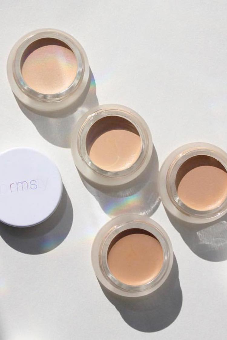 Four RMS Beauty cream pots in varying skin tones on a white surface with rainbow light refractions
