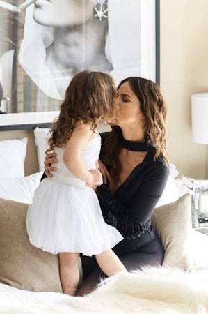 Jodi Gordon kissing her young daughter, who wears a white tutu dress, on a bed at home
