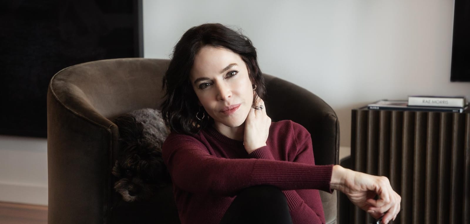 Rae Morris seated in a velvet armchair in a burgundy sweater, hand to her neck, small dog beside her