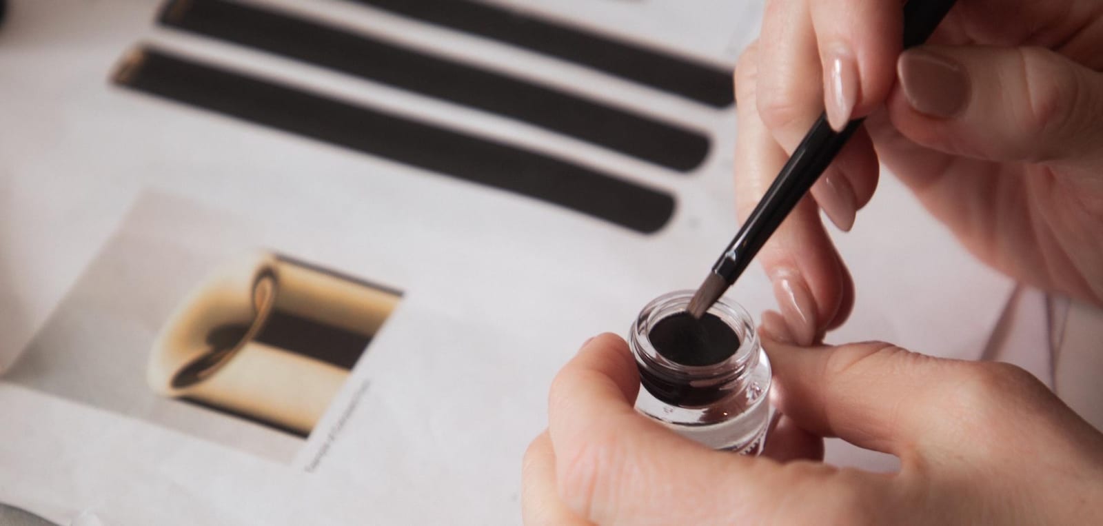 Close-up of hands dipping a fine makeup brush into a small pot of dark gel liner
