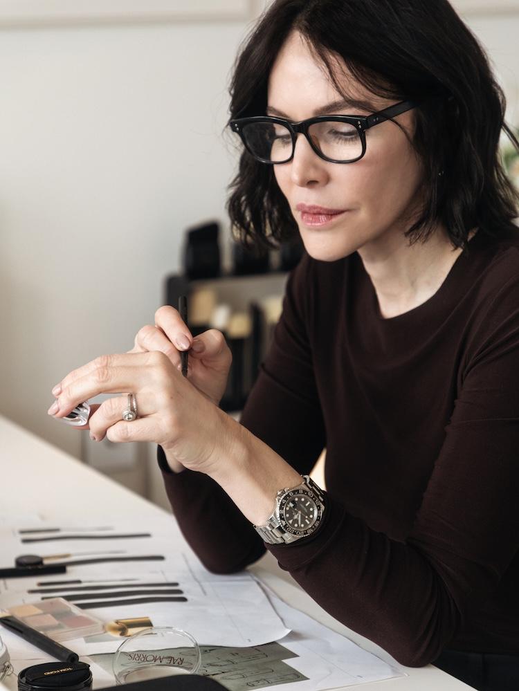 Rae standing with hand in her hair, black blazer over a burgundy tee, glasses, styled bookshelf at left