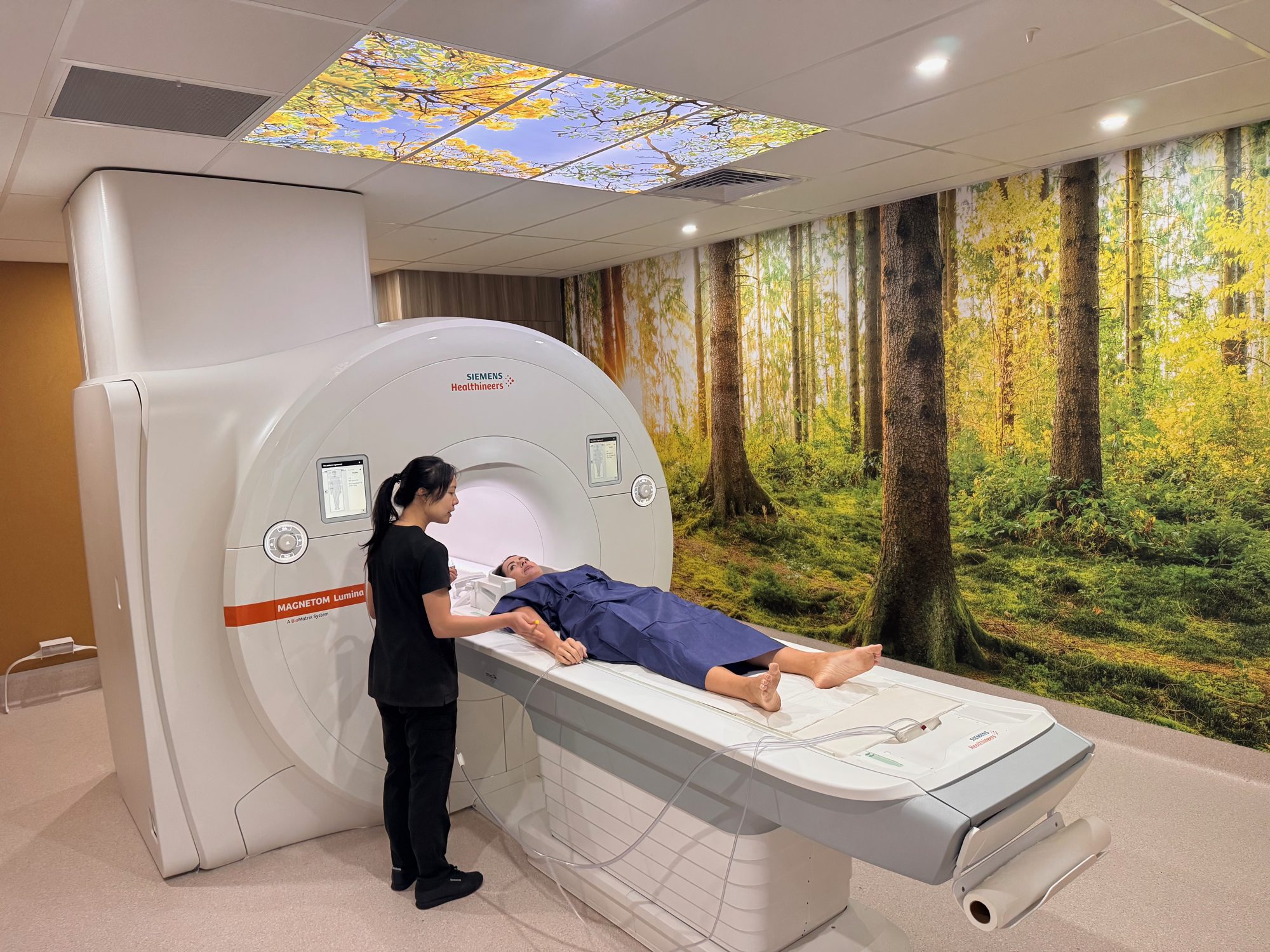 A patient lying on the bed of an MRI scanner while a radiographer prepares the scan, in a room with a forest mural