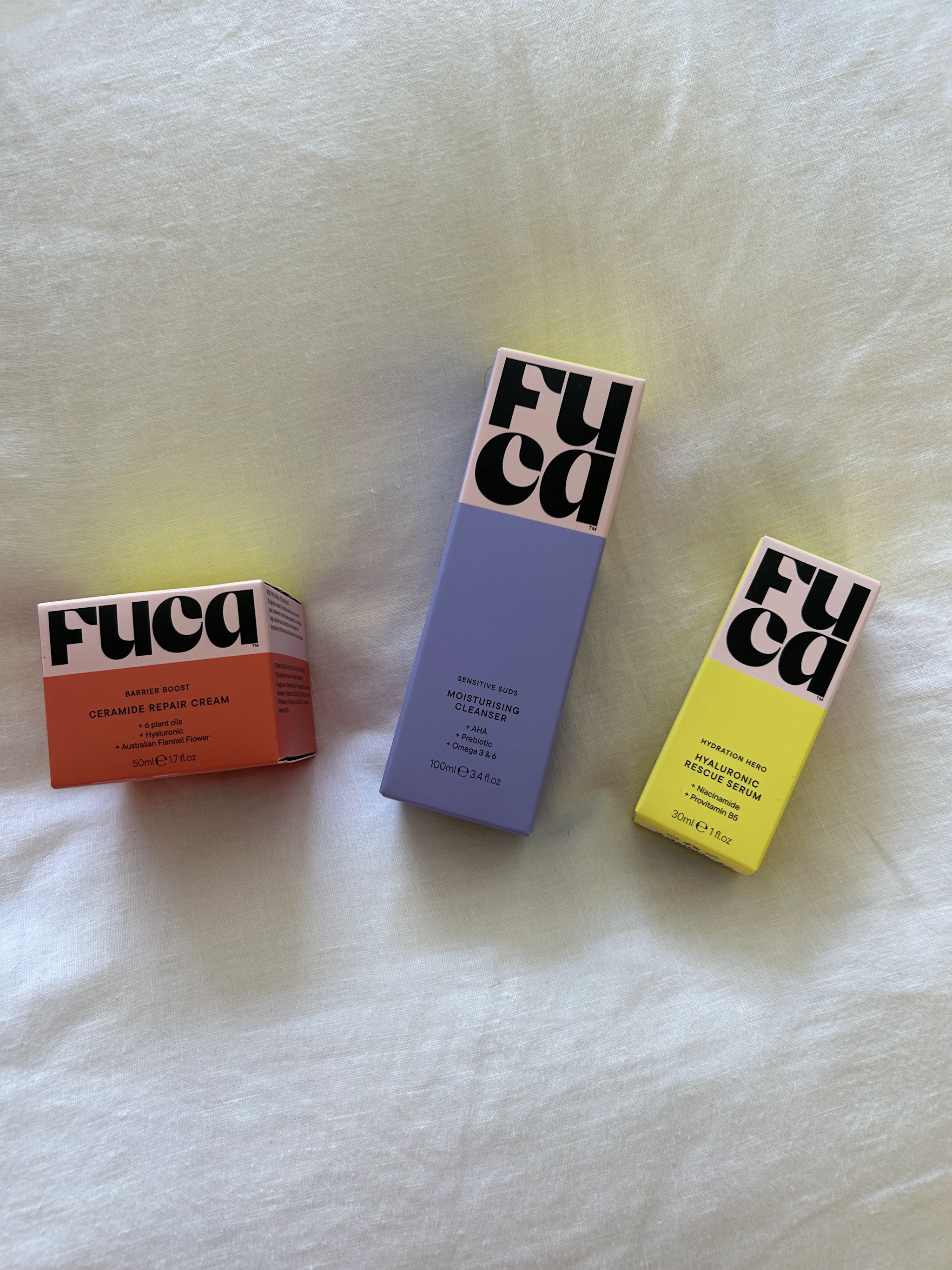 The FUCA skincare range — Ceramide Repair Cream, Moisturising Cleanser and Hyaluronic Rescue Serum — laid out on white linen