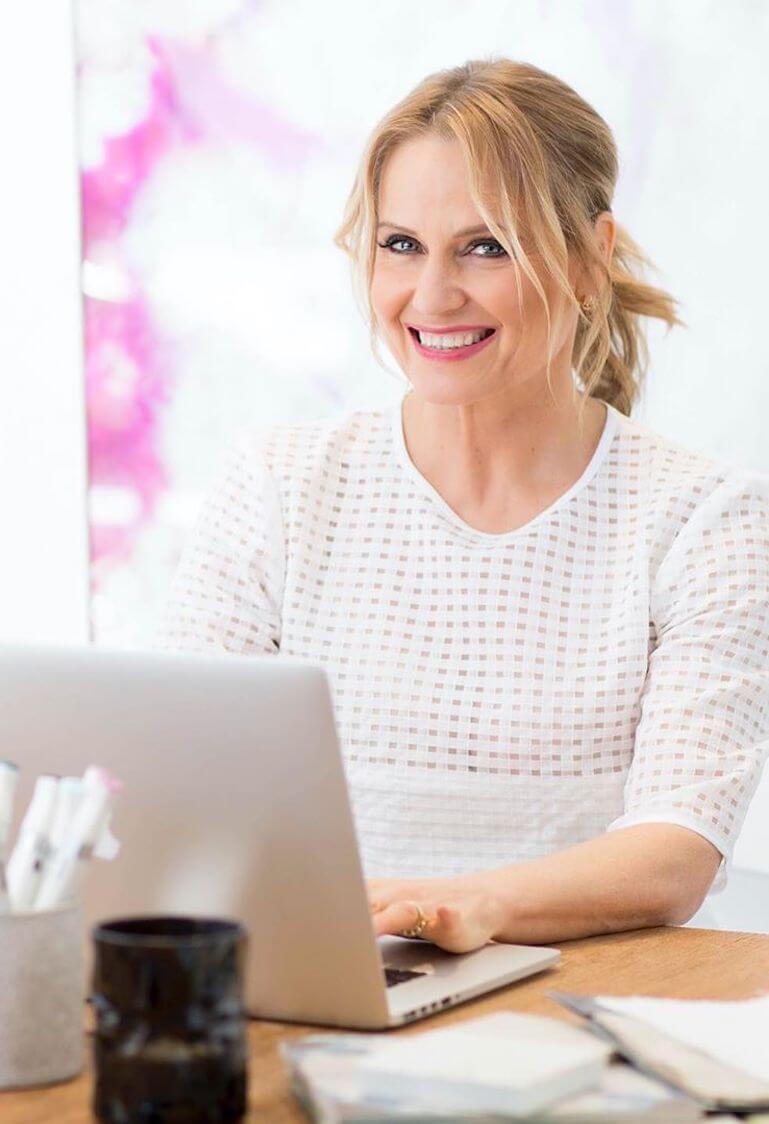 Shaynna Blaze smiling at her desk with a laptop, wearing a sheer check blouse with pink artwork behind